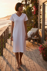 Woman in a white dress standing on a wooden deck by the beach at sunset.